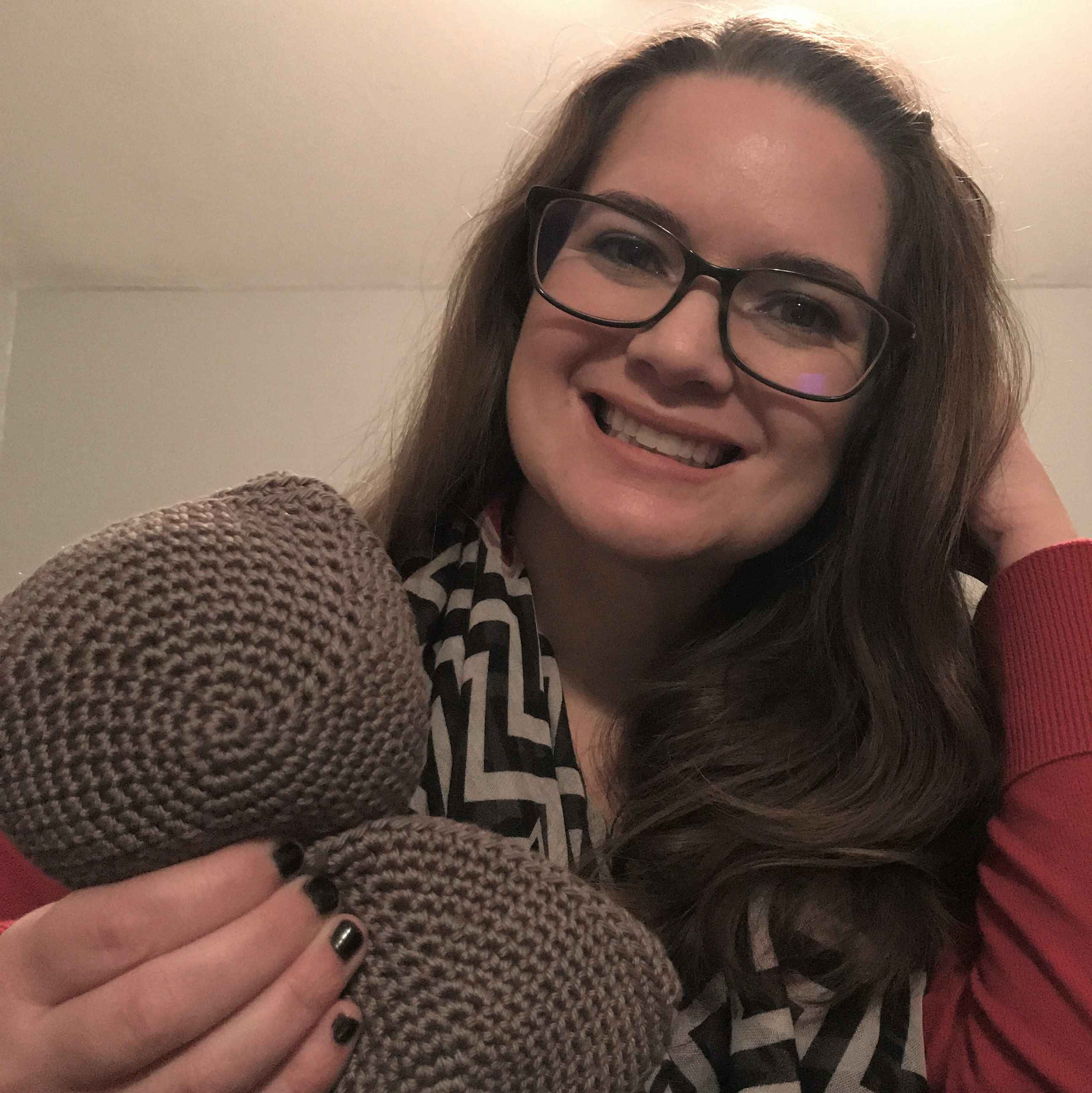 woman with glasses, a red sweater, and black and white cheveron scarf holding a crochet project